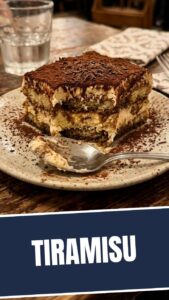 A serving of classic tiramisu in a clear glass dish, showing layers of cream and coffee-soaked ladyfingers